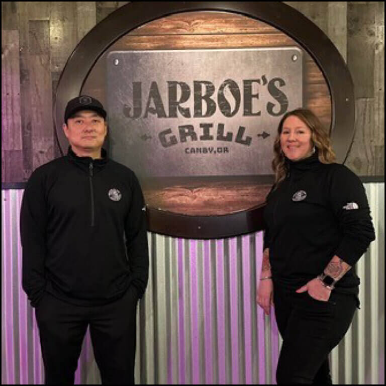 A corrugated metal wall is a backdrop to a wood and metal sign that reads Jarboe's Grill, Canby, Oregon. In front of these signs are a man and woman, standing casually and smiling at the camera.