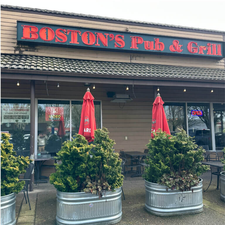 Exterior photo of a restaurant with brown siding and a large red sign that reads Boston's Pub & Grill. In front of the building are several metal tables and chairs, some with red sun umbrellas. In front of all of that are corrugated metal tubs planted with large green shrubs