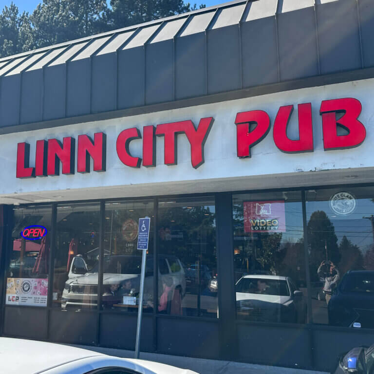 an exterior photo of a large-windowed building in a strip mall setting. There's a Video Lottery sign in the window and the building has a sign with large red letters that reads Linn City Pub