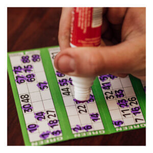A zoomed-in photograph of a paper bingo card, marked with purple spots stamping over an assortment of numbers. A hand hovers over the boar with a bingo marker in hand